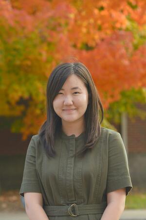 A photo of Jacqueline Yi. She is smiling at the camera and wearing a green shirt. Behind her are autumn trees.