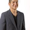 An image of Eric Tang. He is visible from the waist up, and wears a gray blazer over a black shirt. His hair is cut short. He smiles at the camera.