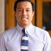 A photo of Dr. Leland Tabares. He is smiling at the camera, and wears a blue shirt and striped blue and white tie.